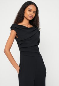 Μαύρο αμάνικο τοπ με ντεκολτέ που πέφτει και απαλό, υφασμάτινο ύφασμα με υφή, με διακριτικές πιέτες και εφαρμοστή σιλουέτα.