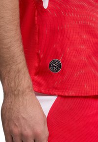 Rotes Sporttrikot mit strukturiertem Stoff und dynamischen, helleren Wellenmustern. Enthält ein rundes Logo, das am unteren Saum aufgenäht ist.