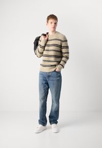 Bežový pruhovaný sveter s čiernymi pruhmi, svetlomodré roztrhané džínsy a biele tenisky; model drží čierny batoh cez rameno.