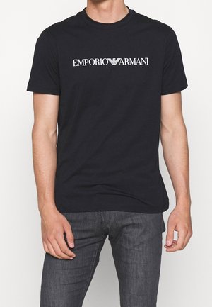 Mann trägt ein schwarzes T-Shirt mit kurzen Ärmeln und weißem "EMPORIO ARMANI" Logo sowie dunkelgraue Jeans, steht vor einfachem weißem Hintergrund.