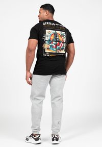 Juodas medvilnės marškinėliai su spalvinga grafika, vaizduojančia gorilą, tropinius elementus ir užrašu "KOL KAS MOTYVUOTIEMS" ant nugaros. Pilkos treniruočių kelnės.