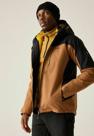 Mann trägt eine braun-schwarze Kapuzenjacke für draußen über einem gelben Reißverschluss-Pullover und einer schwarzen Mütze, posiert mit einer Hand in der Tasche.