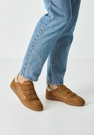 Brązowe zamszowe sneakersy z panelami z siateczki, wyposażone w trzy paski na rzepy i gumową podeszwę, zestawione z niebieskimi dżinsami o prostych nogawkach.