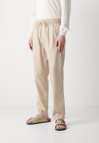 Beige, lette, elastiske bukser med avslappet passform, kombinert med brune sandaler med åpen tå som har et bredt stroppdesign.