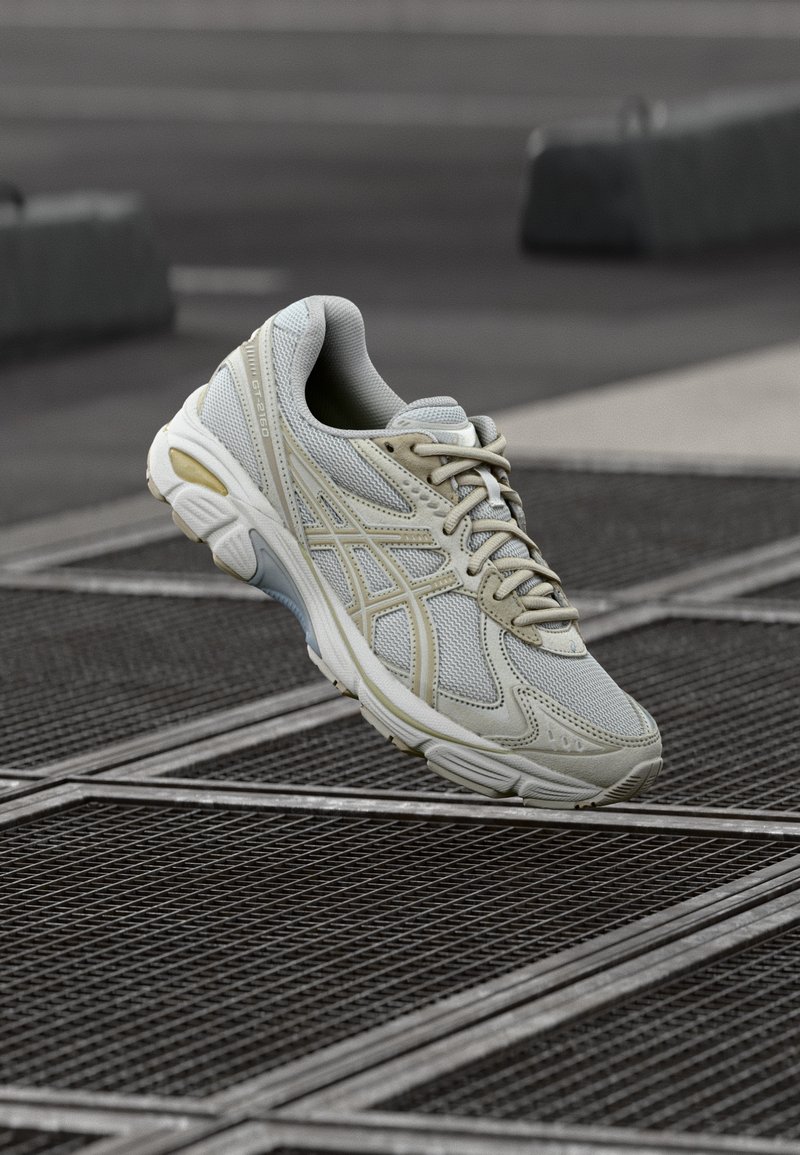 Chaussure de sport gris clair et beige suspendue au-dessus d'une grille métallique sur un sol urbain sombre.