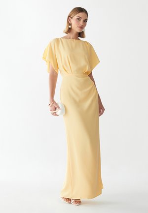 Mujer con un vestido largo hasta el suelo de color amarillo pálido con mangas de vuelo, sostiene un pequeño bolso de mano blanco y lleva sandalias blancas.