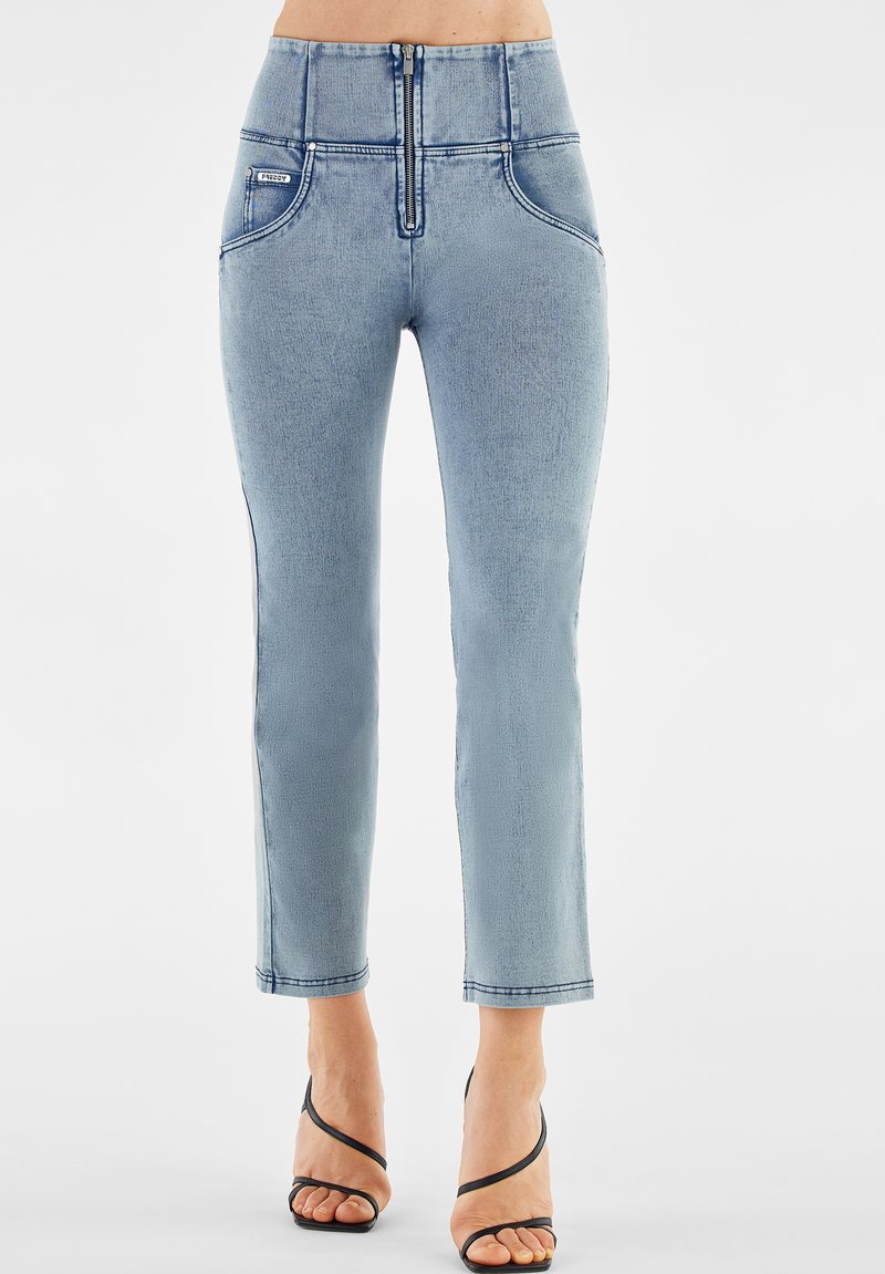 Femme portant un jean slim taille haute bleu clair avec fermeture éclair sur le devant et des sandales à talons hauts noires à lanières, se tenant sur un fond blanc.