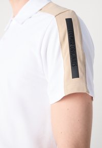 Balta polo marškinių su smėlio spalvos peties juostele, kurioje juodai užrašyta "COLMAR ORIGINALS". Sklandus audinys su klasikiniu apykaklės dizainu.