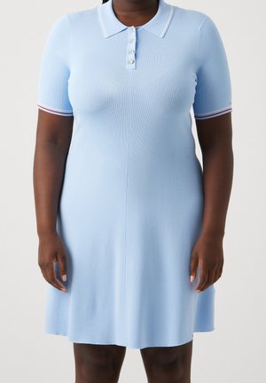 Kobieta w jasnoniebieskiej krótkorękawowej dzianinowej sukience z kołnierzykiem i plisą na guziki, z białym i czerwonym wykończeniem na rękawach.