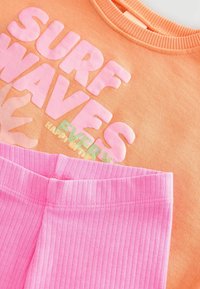 Sudadera naranja con un gráfico en relieve en rosa y verde que dice "SURF WAVES", combinada con leggins rosa de canalé con una cintura elástica.