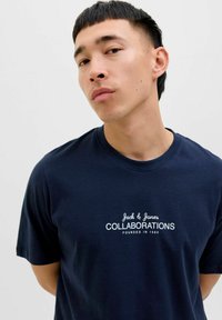 Tumši zils kokvilnas T-krekls ar baltu tekstu: "Jack & Jones Sadarbības dibinātas 1990. gadā" uz krūšu daļas. Apaļš kakla izgriezums, īsas piedurknes.