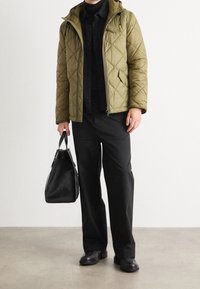 Olivgrüne gesteppte Jacke mit Kapuze, schwarzes Hemd und lockere schwarze Hose; hält eine schwarze strukturierte Handtasche; glatte Lederschuhe.