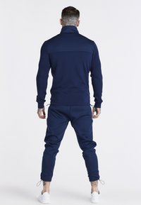 Tuta blu navy con colletto a zip, maniche lunghe e pantaloni affusolati. Presenta polsini elastici e orlo regolabile con cordini.