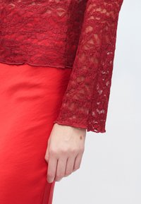 Rdeča čipkasta majica z dolgimi rokavi, z rožnatim vzorcem in valovitim robom. Stilirana z gladko, oprijeto rdečo krilo.