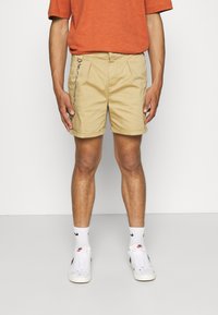 Khaki-Shorts mit lockerer Passform, ausgestattet mit Falten und einem silbernen Kettendetail. Getragen mit weißen Sneakers und gemusterten Socken.