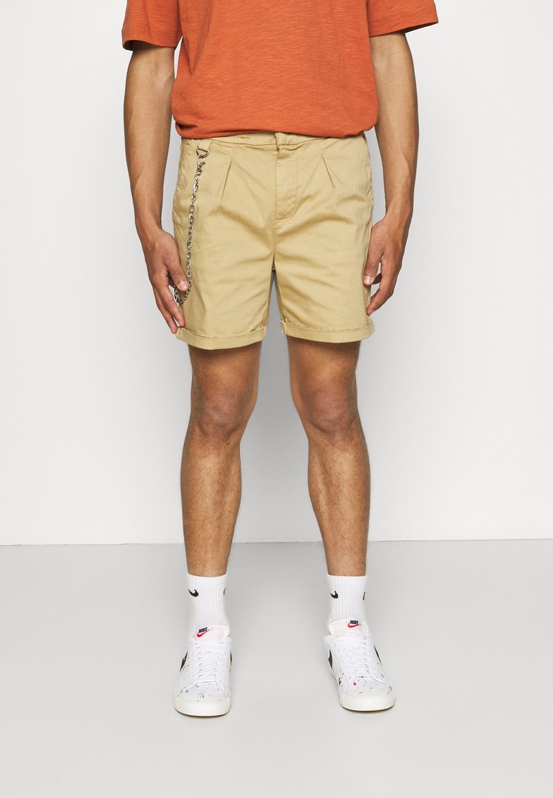 Khaki-Shorts mit lockerer Passform, ausgestattet mit Falten und einem silbernen Kettendetail. Getragen mit weißen Sneakers und gemusterten Socken.