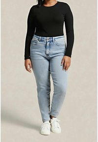 Kvinde iført sort langærmet top, lyseblå højtaljede jeans og hvide sneakers, stående mod en ensfarvet baggrund.