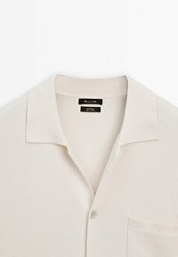 Polo-shirt in lichtbeige stof met een gestructureerde brei, voorzien van een kraag, een enkele borstzak en een sluiting met knopen aan de voorkant. Etiket zichtbaar aan de binnenkant.