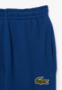 Blauwe katoenen joggers met elastische tailleband, zijzakken en geborduurd groen krokodillenlogo op de linker bovenbeen. Soepele textuur.