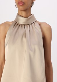 Top halter neck in satin beige con design a plissettature e una superficie liscia, caratterizzato da un colletto alto e spalle scoperte.