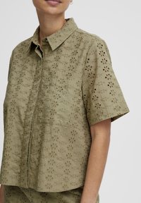 Olive green short-sleeve shirt with floral eyelet pattern, button-down collar, and a loose fit. Textured fabric with subtle detailing.