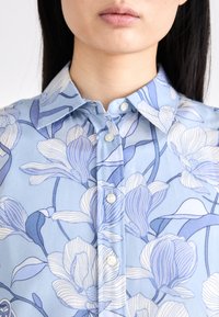 Light blue button-up shirt featuring a floral pattern in various shades of blue and white, with a classic collar and smooth fabric texture.
