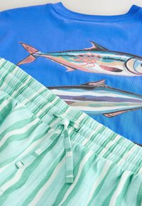 Gros plan sur une chemise bleue avec deux imprimés de poissons colorés, associée à un short à taille élastique rayé vert et blanc avec cordon de serrage.