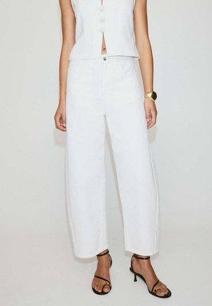 Femme portant un pantalon blanc taille haute à jambes larges, un top blanc sans manches boutonné, des sandales noires à talons et un bracelet en or au poignet droit.
