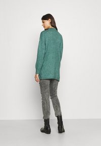 Cardigan tricotat teal cu guler înalt, spate prelungit și manșete cu model, purtat cu pantaloni gri texturat și cizme negre până la glezne.