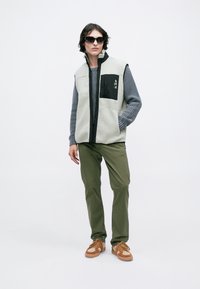 Svijetlosivi flis prsluk s crnim detaljima i prednjim džepom, uparen s sivoj rebrastim džemperom, maslinastim hlačama i smeškim obućom.