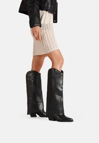 Stivali alti neri in pelle, con una texture liscia e elegante e tacco a blocco, abbinati a una gonna in maglia a coste di colore crema.