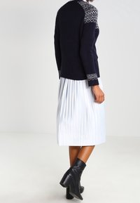Navy-Pullover mit gemustertem Schulterbereich, weißem, plissiertem Rock und schwarzen Knöchelstiefeln. Das Outfit zeichnet sich durch strukturierte Materialien und kontrastierende Farben aus.