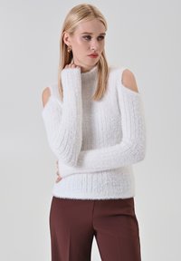 Pull en maille blanche avec design cold-shoulder, fabriqué dans un matériau fluffy. Porté avec un pantalon taille haute marron. Détails minimalistes, texture douce.