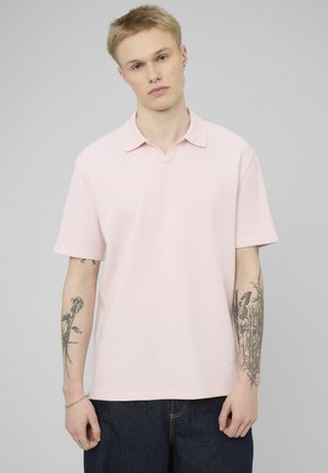 Jeune personne aux cheveux blonds courts portant une chemise à col rose pâle à manches courtes et un jean foncé, montrant des tatouages sur les deux avant-bras.