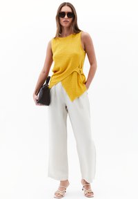 Gul texturerad ärmlös topp med knytdetalj, matchad med elfenbensfärgade vida byxor och vita sandaler med remmar. Modellen bär också solglasögon.