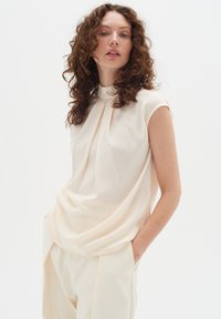 Cream-colored sleeveless top with a high collar and draped design. Soft, smooth fabric with subtle texture. Paired with light-colored trousers.