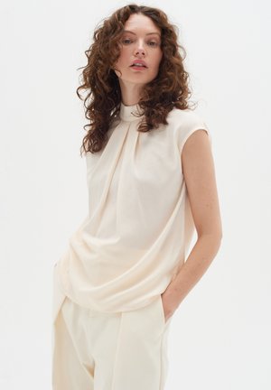 Cream-colored sleeveless top with a high collar and draped design. Soft, smooth fabric with subtle texture. Paired with light-colored trousers.