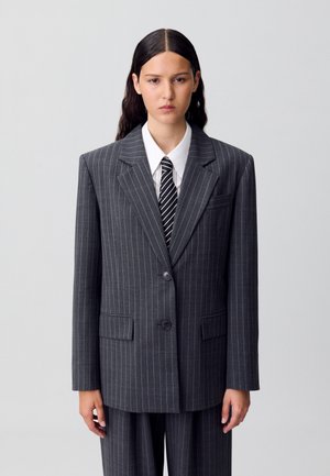 Costume gris à rayures avec une veste structurée à deux boutons et des poches latérales, accompagnée d'une chemise blanche et d'une cravate à rayures noires.