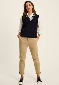 Navy knitted cable vest with a v-neck, layered over a white shirt. Tan rolled-up chinos and dark sneakers complete the outfit.