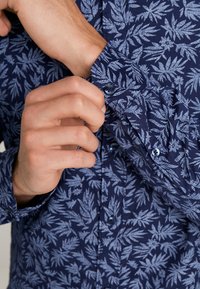 Camicia a maniche lunghe blu navy con un motivo di foglie blu chiaro. Presenta polsini con bottoni e una texture del tessuto liscia. Primo piano del dettaglio del polsino.