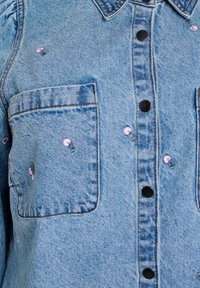 Svetlo modrá denimová košeľa s čiernymi gombíkmi, dvoma prednými vreckami a ružovou kvetinovou výšivkou na látke.