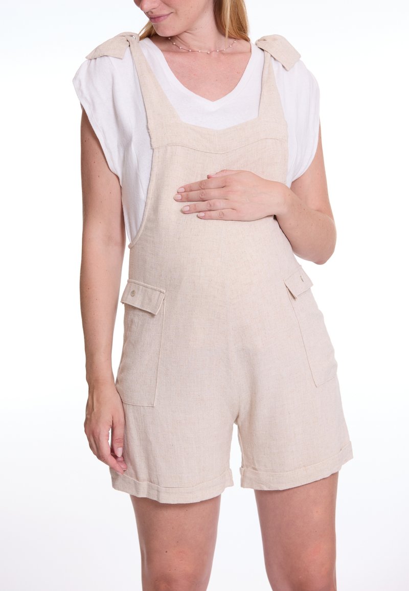 Beige linnen salopettes met een ontspannen pasvorm, voorzien van schouderdetails, grote voorzakken en omgeslagen zoom. Draag ze over een wit T-shirt.