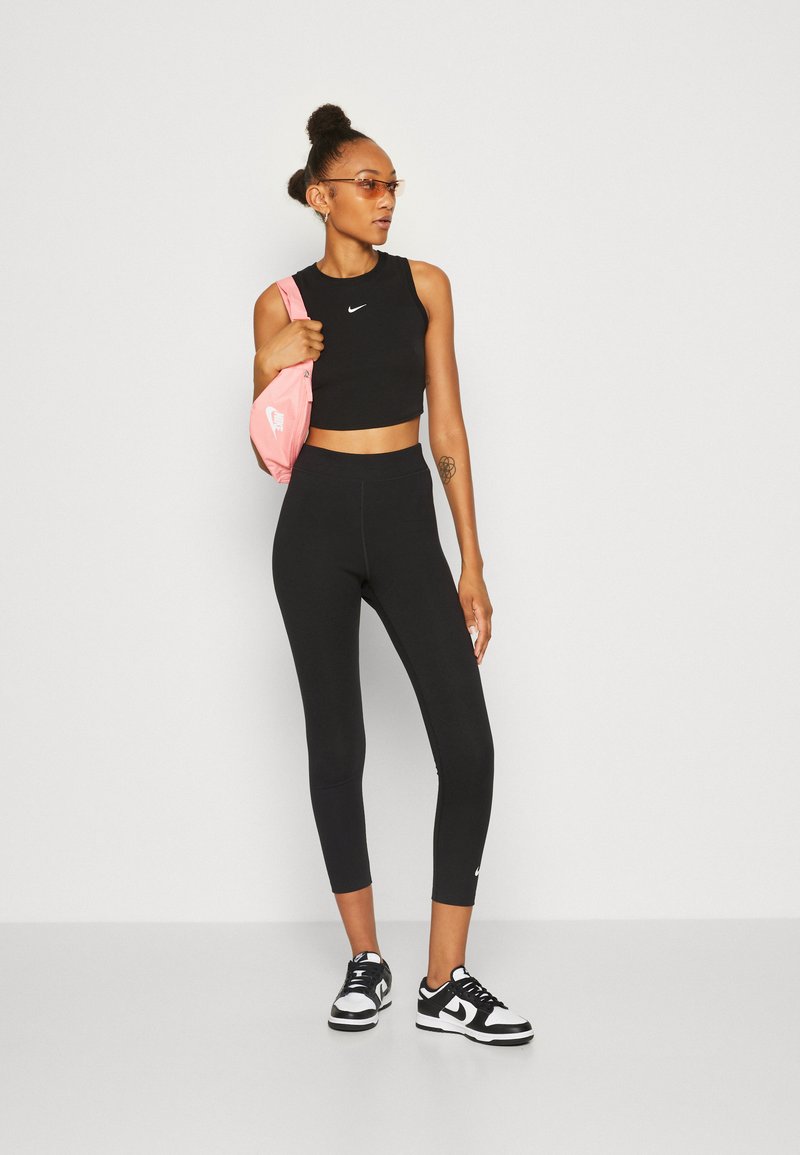 Un crop top de sport noir et un legging taille haute, associés à des baskets noir et blanc. Un sac rose pend à l'épaule. Des lunettes de soleil transparentes sont portées.