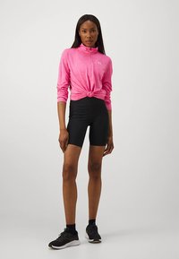 Roze lange mouwen top met ritssluiting, geknoopt in de taille, gecombineerd met zwarte hoge taille fietsbroekjes. Model draagt zwarte sportieve schoenen.