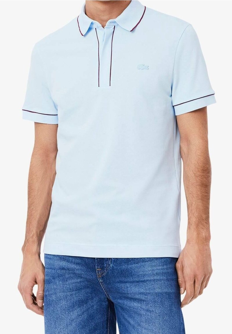 Homme portant un polo bleu clair avec des bordures foncées au col et aux manches, associé à un jean bleu, debout devant un fond blanc.