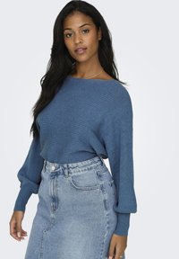 Pull en maille bleu à manches larges, associé à une jupe en denim clair taille haute dotée de poches avant et d'une fermeture à boutons.
