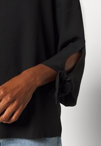 Bras portant un blouse noire avec des manches fendues nouées, et un jean en denim bleu clair visible contre un fond blanc.