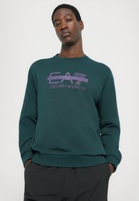 Sweatshirt verde com decote redondo, apresentando um logótipo roxo da Emporio Armani no peito. Tecido suave e texturizado, corte descontraído.