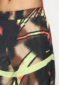 Primer plano de la cintura de una persona que lleva puestos unos pantalones cortos negros con un patrón abstracto en marrón, beige, verde neón y naranja, y un pequeño logo de Nike en color verde.