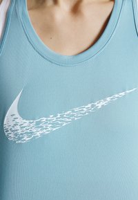 Gros plan d’une personne portant un haut sans manches bleu clair avec un grand logo Nike Swoosh blanc à design texturé.